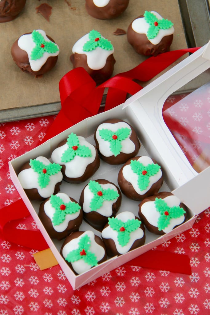 Nine truffles with fondant holly on top, in a white box. there are 5 other truffles above the box on a tray.
