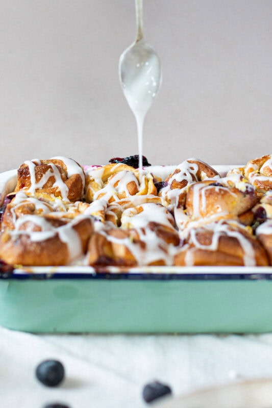 A side shot of a spoon drizzling icing on lemon and blueberry rolls
