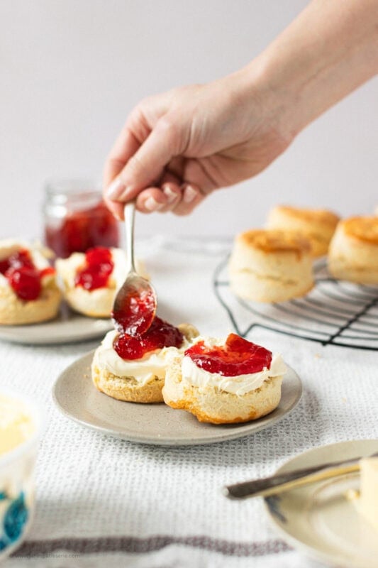 A hand spooning jam onto a buttered classic buttery scone